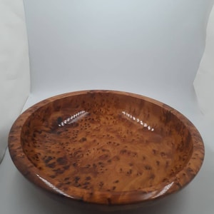 May include: A round wooden bowl with a polished, glossy finish. The bowl is crafted from light brown wood, featuring darker brown patterns. The bowl's smooth surface and wide opening make it suitable for serving or display. The diameter is approximately 25 cm.