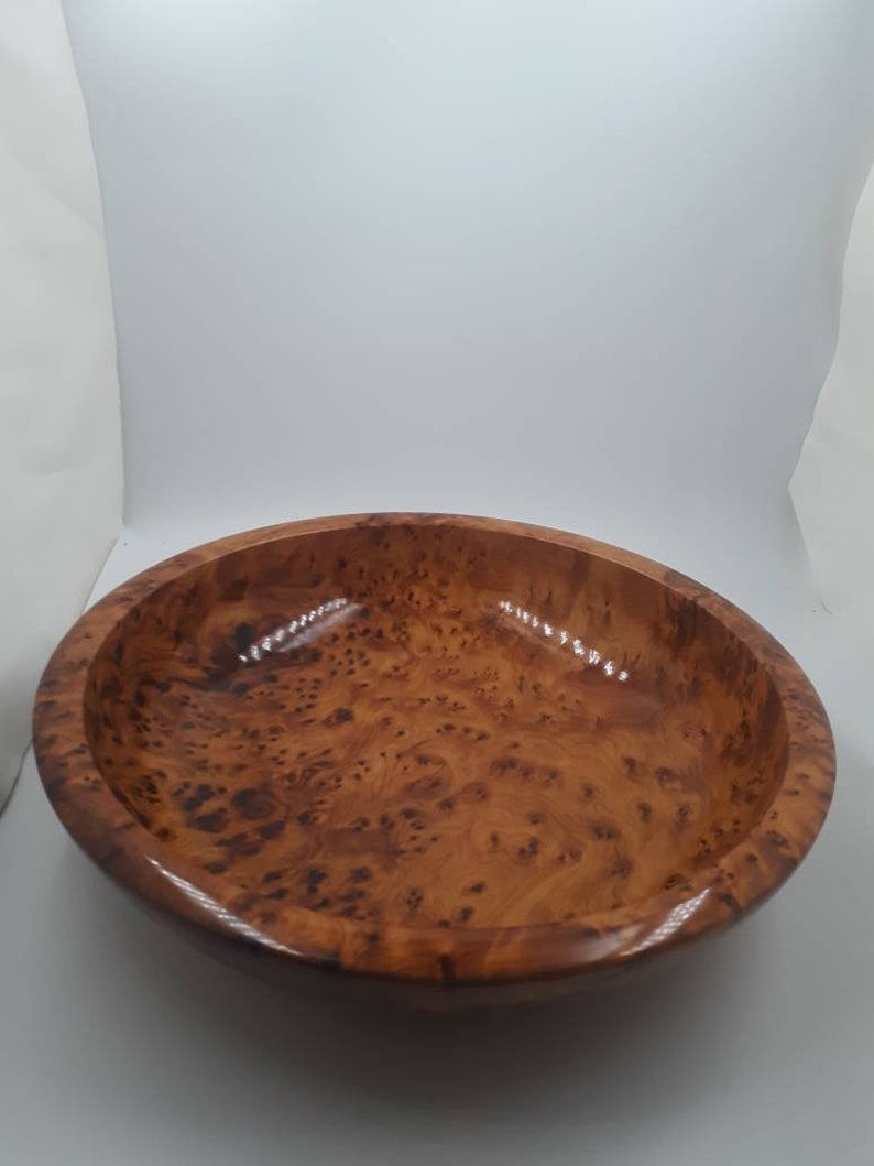 May include: A round wooden bowl with a polished, glossy finish. The bowl is crafted from light brown wood, featuring darker brown patterns. The bowl's smooth surface and wide opening make it suitable for serving or display. The diameter is approximately 25 cm.