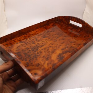 May include: A rectangular wooden serving tray with a rich, warm brown burl wood grain pattern. The tray has curved sides with cut-out handles and a glossy finish. The tray is being held by a hand.