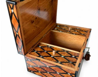 Puede incluir: Una caja de madera con la tapa abierta que presenta un acabado de madera veteada marrón y un patrón de rombos negros incrustado con pequeños detalles blancos. El interior contiene una bandeja extraíble a juego y una borla marrón en el lateral.