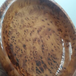 May include: Oval wooden bowl with a glossy finish. The bowl has a natural wood grain pattern in shades of brown and tan. The interior of the bowl is smooth and polished, reflecting light. The bowl is likely used for serving or display.
