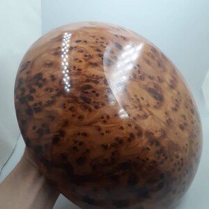 May include: A large, oval wooden bowl with a polished surface. The bowl is a warm brown hue, displaying a distinctive burl wood grain pattern with dark spots and swirling textures. The bowl is held by a person's hand.