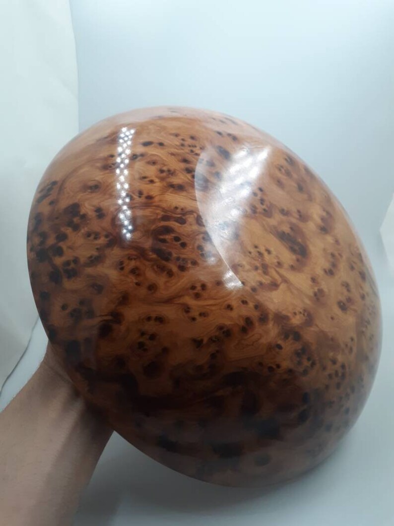 May include: A large, oval wooden bowl with a polished surface. The bowl is a warm brown hue, displaying a distinctive burl wood grain pattern with dark spots and swirling textures. The bowl is held by a person's hand.