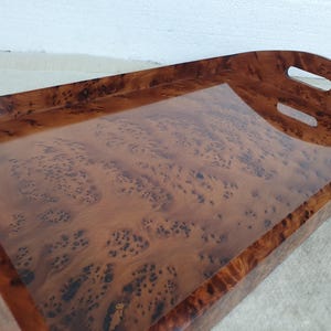 May include: A rectangular wooden serving tray with a glossy finish. The tray has a rich, brown burl wood pattern and two handle cutouts. The tray is designed for serving food or drinks.