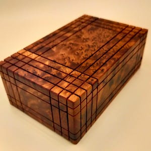 Può includere: Una scatola di legno con un design geometrico intricato. La scatola è realizzata in legno scuro con una tonalità rossastra e ha una finitura liscia e lucida.