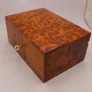 Peut inclure: Une boîte en bois avec un trou de serrure et une clé dorée. La boîte est en bois brun clair avec un motif de grain brun foncé.