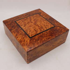 Peut inclure: Une boîte carrée en bois avec un placage en bois de ronce et une incrustation noire. La boîte a un couvercle à charnières et est faite de bois de haute qualité.