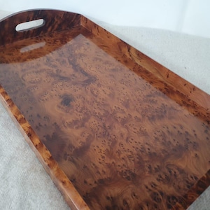 May include: A rectangular wooden serving tray with a glossy finish. The tray features a rich, warm brown burl wood grain pattern. It has a curved handle with a cutout for easy carrying.