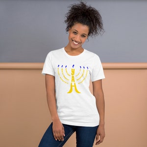May include: White t-shirt with a gold menorah design. The menorah is made up of the word "YAH" with the Hebrew words for "strength, forgiveness, compassion, kindness, peace, truth, and everlasting love" surrounding it.