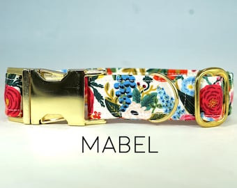 May include: A white dog collar with a floral print featuring red, blue and yellow flowers. The collar has a gold metal buckle and D-ring. The text "MABEL" is printed below the collar.