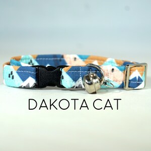 Könnte beinhalten: Ein blaues, oranges und weißes Katzenhalsband mit einer schwarzen Schnalle und einer silbernen Glocke. Das Halsband trägt den Text "DAKOTA CAT".