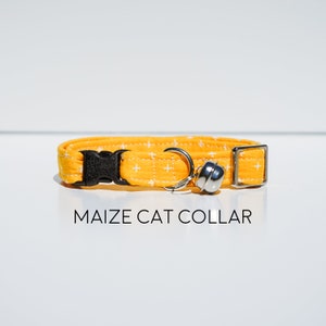 May include: A yellow cat collar with a white cross pattern and a silver bell. The collar has a black plastic buckle.