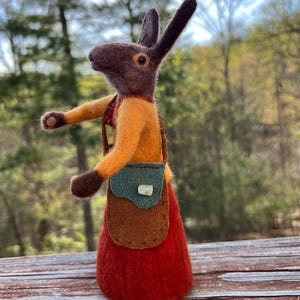 Peut inclure: Un lapin en feutre brun portant une chemise jaune, une jupe brune et un sac en cuir brun avec un rabat vert et un bouton blanc. Le lapin est debout sur une surface en bois.