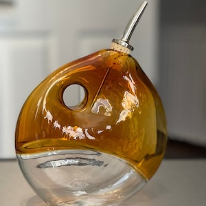 May include: A clear glass bottle with a brown amber top and a silver spout. The bottle has a unique, rounded shape with a hole in the side.
