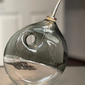 May include: A clear glass bottle with a grey tint and a silver spout. The bottle has a unique design with a hole in the side.
