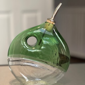 May include: A green glass bottle with a clear base and a silver spout. The bottle is round in shape with a hole in the side.