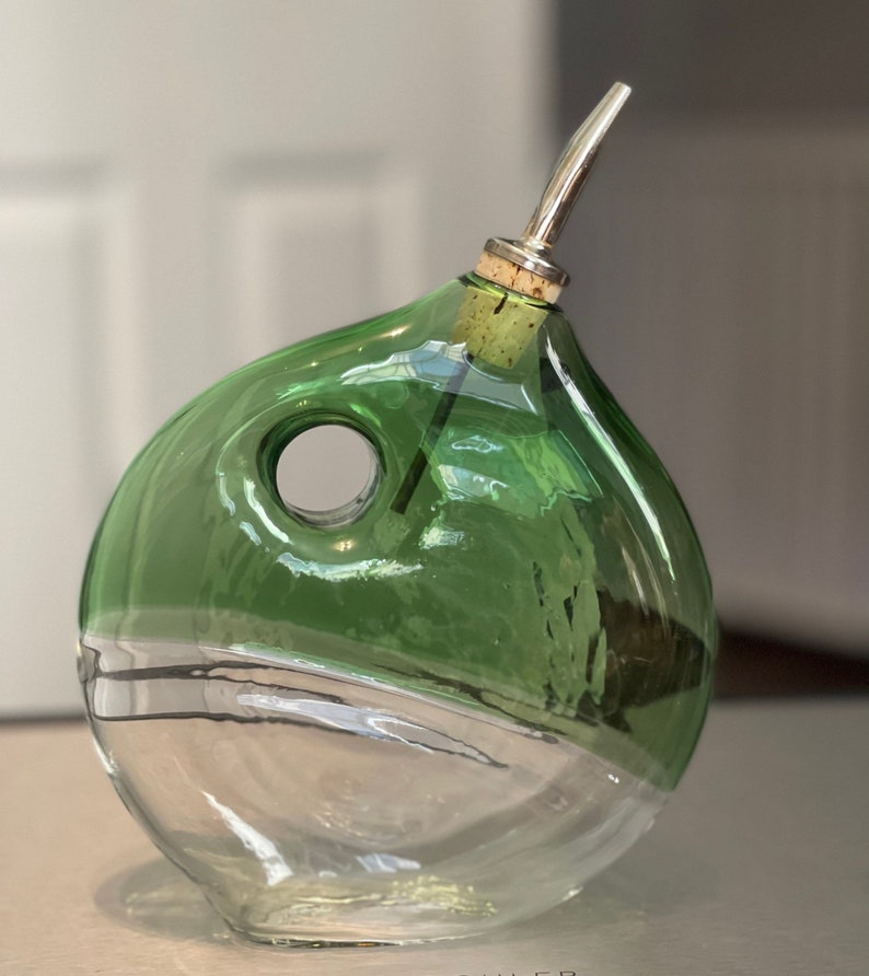 May include: A green glass bottle with a clear base and a silver spout. The bottle is round in shape with a hole in the side.