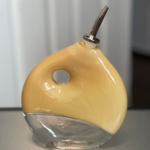 May include: A yellow glass bottle with a clear glass base and a silver pouring spout. The bottle has a unique shape with a hole in the side.