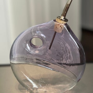 May include: A clear glass bottle with a purple tint and a cork stopper. The bottle has a unique shape with a hole in the side. It is designed for pouring liquids.