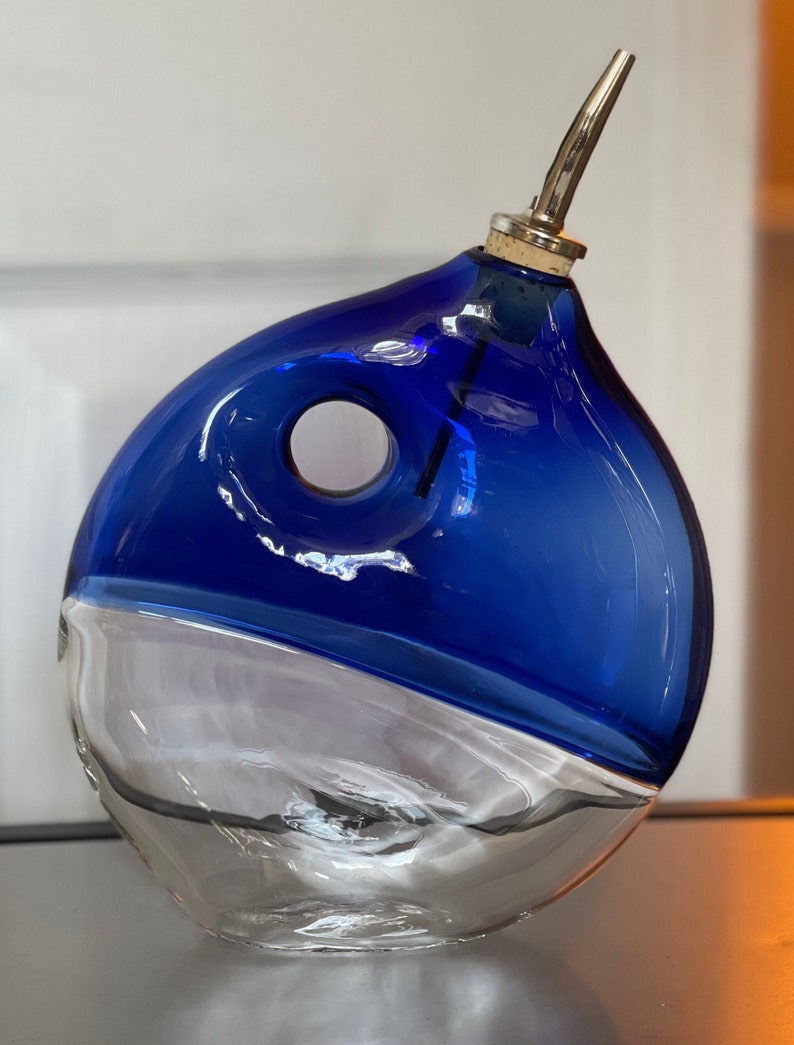 May include: A blue and clear glass oil and vinegar bottle with a spout. The bottle has a unique, rounded shape with a hole in the side.