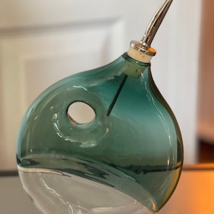May include: A teal blue glass bottle with a clear glass base and a silver pouring spout. The bottle has a unique shape with a hole in the side.