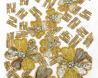 May include: A white fabric background with gold and silver beaded flowers. The flowers are made of sequins and beads and have a 3D effect.