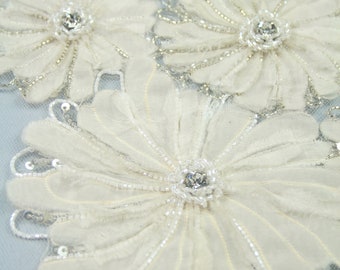 May include: Close-up of white fabric flowers with beaded edges and rhinestone centers. The flowers are arranged on a light blue background.
