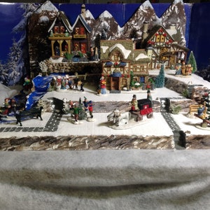 May include: A miniature Christmas village scene with houses, trees, and figures. The village is set on a snowy base with a blue river flowing through it. There are several figures of people and animals, including a horse-drawn carriage.