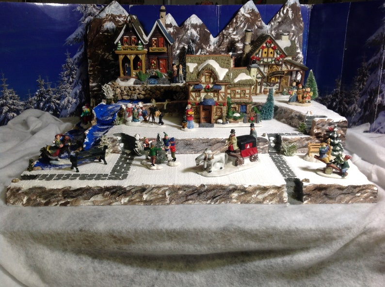 May include: A miniature Christmas village scene with houses, trees, and figures. The village is set on a snowy base with a blue river flowing through it. There are several figures of people and animals, including a horse-drawn carriage.
