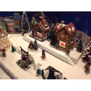May include: A miniature Christmas village scene with two lit houses, a small tree, and figurines. The houses are decorated with red roofs and white trim. The scene is set on a white base with a snowy effect.