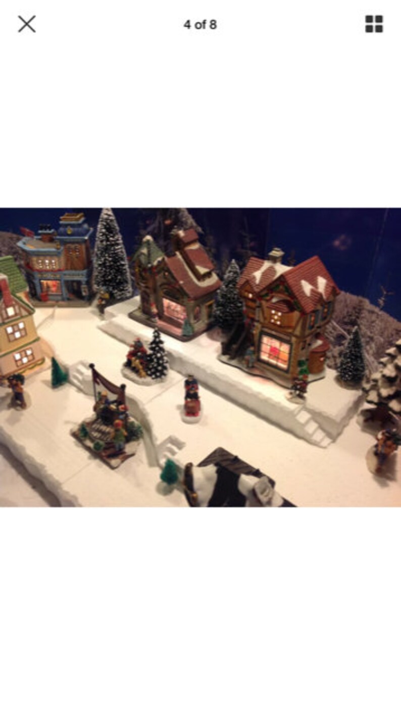 May include: A miniature Christmas village scene with two lit houses, a small tree, and figurines. The houses are decorated with red roofs and white trim. The scene is set on a white base with a snowy effect.