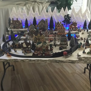 May include: A miniature Christmas village scene with a train track, snow-covered houses, and a snowy mountain backdrop. The village is lit with blue lights and features a variety of buildings, including a church, a bakery, and a toy store.
