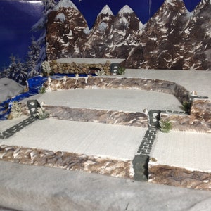 May include: A snowy mountain scene diorama with a blue river, stone paths, and small trees. The diorama is made of cardboard and painted to look like a winter landscape.