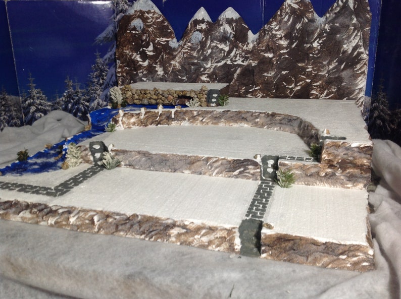 May include: A snowy mountain scene diorama with a blue river, stone paths, and small trees. The diorama is made of cardboard and painted to look like a winter landscape.