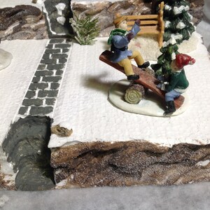 May include: Two children on a wooden seesaw in a snowy winter village scene. The seesaw is made of a log with a wooden plank on top. The children are wearing winter clothes and are smiling. There is a small evergreen tree and a wooden bench in the background.