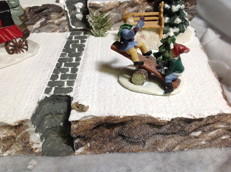 May include: Two children on a wooden seesaw in a snowy winter village scene. The seesaw is made of a log with a wooden plank on top. The children are wearing winter clothes and are smiling. There is a small evergreen tree and a wooden bench in the background.