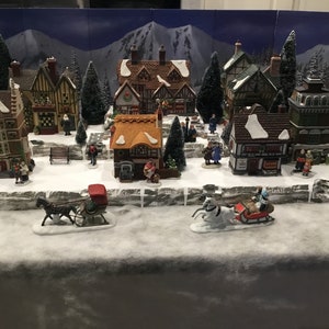 May include: A miniature Christmas village scene with snow-covered houses, trees, and figures. The village is set on a snowy base with a stone wall and includes two horse-drawn sleighs.