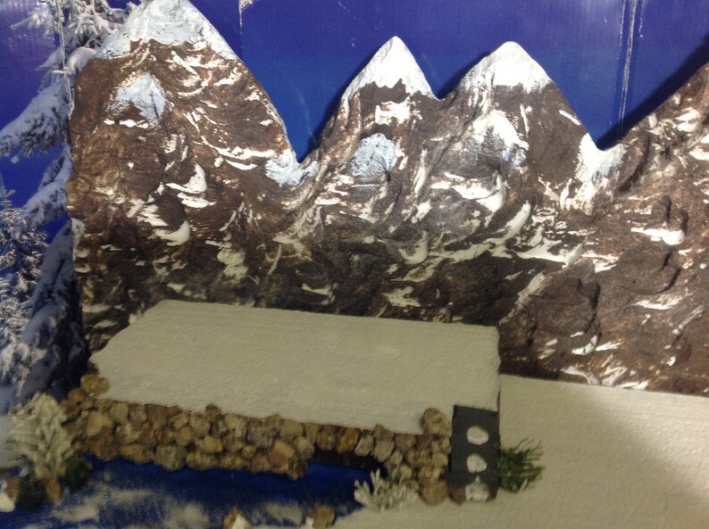 May include: A miniature winter scene with a snowy mountain range backdrop, a small stone wall, and a white snowy surface in the foreground.