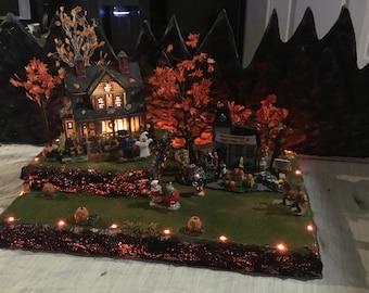 May include: A miniature Halloween village scene with a house, trees, and figures. The scene is lit with orange lights and features a sign that reads "Halloween Festival 2019".