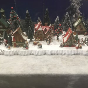 May include: A miniature Christmas village scene with a church, houses, and trees covered in snow. The village is lit up with lights and features small figurines of people and animals.
