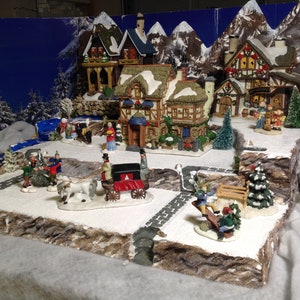 May include: A miniature Christmas village scene with snow-covered houses, trees, and figures. The village is set against a backdrop of blue mountains and a snowy landscape.