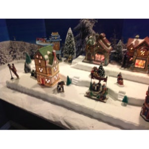 May include: A miniature Christmas village scene with a variety of houses, trees, and figures. The houses are lit up with white lights and there is a small horse and carriage in the foreground.