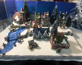 Puede incluir: Una escena de un pueblo navideño en miniatura con casas, árboles y un río cubiertos de nieve. El pueblo presenta una iglesia, tiendas y casas, todas decoradas para la temporada navideña. El telón de fondo representa un bosque nevado.