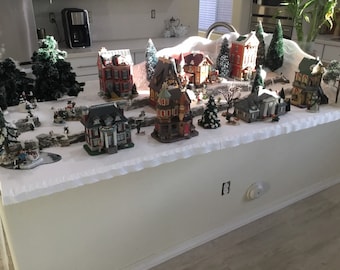 May include: A miniature Christmas village scene with snow-covered houses, trees, and a white tablecloth. The houses are decorated with lights and have a festive feel.