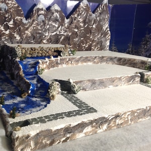 May include: A miniature winter scene with a blue river flowing through a snowy landscape. The scene features a backdrop of brown and white mountains, a stone bridge, and a small stone path.