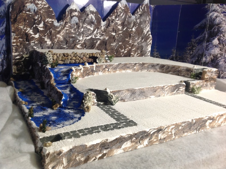 May include: A miniature winter scene with a blue river flowing through a snowy landscape. The scene features a backdrop of brown and white mountains, a stone bridge, and a small stone path.
