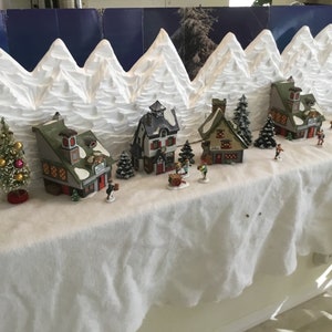 May include: A miniature Christmas village scene with white styrofoam mountains, green trees, and small houses with red and white accents. The houses are decorated with snow and lights.