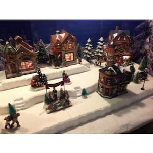 May include: A miniature Christmas village scene with several houses, trees, and figures. The houses are decorated with lights and have snow-covered roofs. The scene is set on a white base with a snowy landscape.