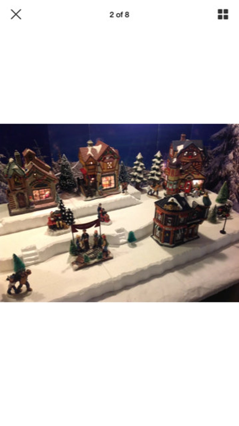 May include: A miniature Christmas village scene with several houses, trees, and figures. The houses are decorated with lights and have snow-covered roofs. The scene is set on a white base with a snowy landscape.