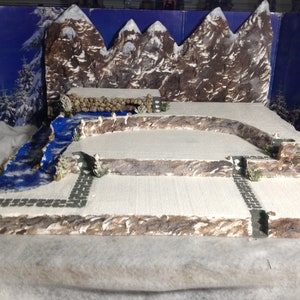 May include: A miniature winter scene with a blue river, snow-covered ground, and a backdrop of brown mountains. The scene is built on a multi-level platform with stone walls and paths.